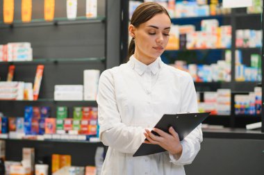 Deneyimli beyaz önlüklü kadın eczacı envanterleri kontrol ediyor, modern eczanede elinde ilaçlı raflar arasında bir pano tutuyor.