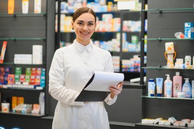 Gülümseyen kimyager laboratuvar önlüğü giyiyor ve elinde bir klip var. Rafların önünde duruyor. Sağlık ürünleri ve ilaçlarla dolu.