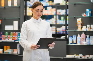 Eczacı aşağıya bakıyor, elinde bir pano ve kalem, modern bir eczanede raflardaki stokları ve ürünleri kontrol ediyor.