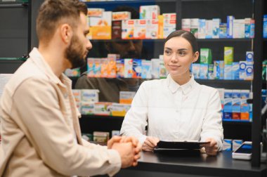 Kadın eczacı, reçeteleri tartışmak ve modern bir eczanede profesyonel sağlık hizmetleri tavsiyesi vermek için tezgahta erkek müşteriye yardım ediyor.