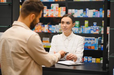 Dispanser tezgahında duran eczacı, bir müşteriye ilaç alımı ve sağlık hizmeti sağlamada yardımcı oluyor.