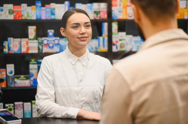 Genç bayan eczacı tezgahtaki bir erkek müşteriyle gülümsüyor ve etkileşime geçiyor, arkasında ilaç rafları var.