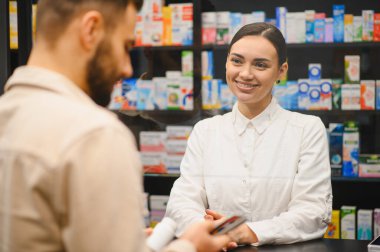 Dost canlısı kadın eczacı, erkek müşteriye resepsiyonda yardım ediyor, modern bir eczanede sağlık hizmeti ve konsültasyon sağlıyor.