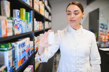 Genç kadın eczacı ya da eczacı bir eczane koridorunda duruyor, bulanık raflara karşı beyaz bir ilaç şişesi tutuyor.