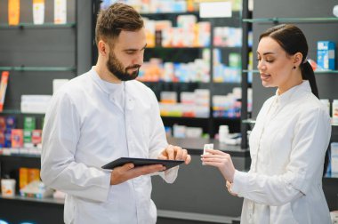 Beyaz önlüklü eczacılar envanter ve hasta bilgileri için dijital tablet kullanırken, ilaç paketlerini tartışırken, modern eczane perakendecisi