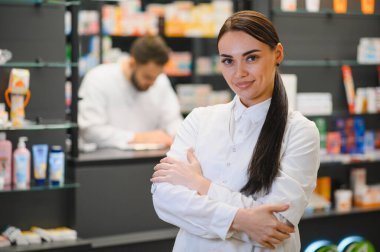 Modern eczanede kolları bağlı duran eczacı profesyonel sağlık hizmeti sunuyor. Arka planda çalışan erkek arkadaş