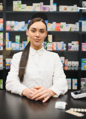 Modern bir eczanede çalışan genç bir kadın eczacı, kameraya bakıyor ve müşterilere ilaçla yardım ederken gülümsüyor.