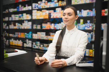 Kasanın başında duran genç bir kadın eczacı elinde ilaç şişesi ve kalemle arka planda ilaç rafları var.