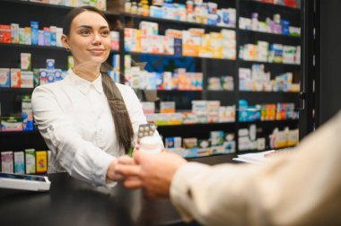 Gülümseyen eczacı, modern bir eczanenin tezgahındaki müşteriye su toplayan paketler ve bir şişe ilaç veriyor.