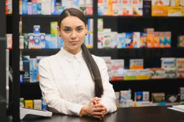Beyaz önlüklü kadın eczacı, kameraya bakıp gülümsüyor, modern bir eczanede raflarda ilaçlarla bekliyor.