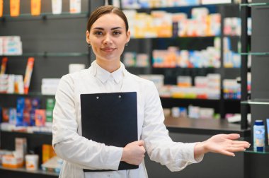 Gülümseyen eczacı kadın elinde bir pano, modern bir eczanede raflarda tıbbi ve kozmetik ürünler sunuyor.