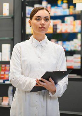 Üniformalı bir eczacı, elinde ilaç raflarıyla modern bir eczanede bir pano tutuyor, envanter ve sağlık ürünlerini yönetiyor.