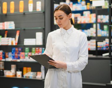 Beyaz üniformalı bir kadın eczacı modern bir eczanede ilaç raflarıyla envanter listesini kontrol ediyor.