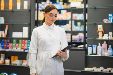 Eczanede elinde panoyla duran genç bir kadın eczacı raflardaki tıbbi malzeme ve ürünlerin envanterini kontrol ediyor.