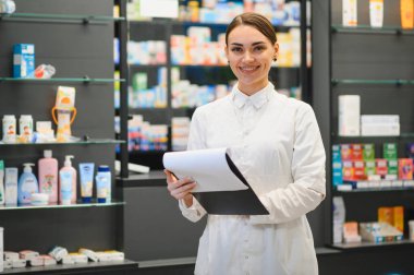 Beyaz önlüklü gülümseyen eczacı modern bir eczanede envanter kontrolü yapıyor ve raflarda ilaç ve ürünler var. İş yerinde profesyonel sağlık hizmeti
