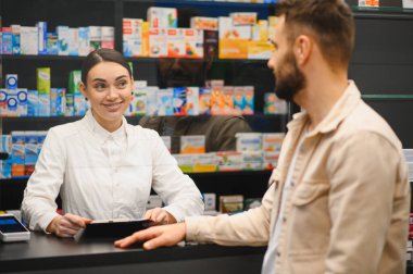 Eczacı gülümsüyor, elinde bir pano tutarken perakende satış yapan bir eczanede erkek müşteriyle konuşuyor.
