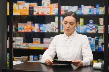 Tezgahta duran genç kadın eczacı arka planda çeşitli ilaçlarla dolu rafları analiz ediyor.