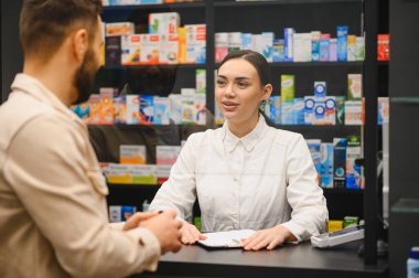 Camın arkasındaki tezgahta duran eczacı, erkek bir müşteriyle ilaç ve reçeteler hakkında konuşuyor.