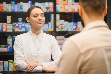 Kadın eczacı tezgahın önünde duruyor, gülümsüyor ve modern bir eczane ortamında müşteriyle konuşuyor.
