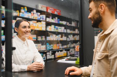Eczacı gülümsüyor, ilaç şişesini tutuyor ve koruyucu ekranın arkasında bir erkek müşteriyle konuşuyor.