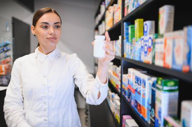 Modern bir eczanede çalışan eczacı kadın, elinde ilaç şişesiyle raflardaki ürünleri kontrol ediyor, sağlık hizmetleri sunuyor.