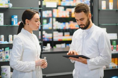 Profesyonel bir eczanedeki dijital tabletin ilaç bilgilerini tartışan ve kontrol eden eczacılar, uzman sağlık hizmetleri sunmaktadır.