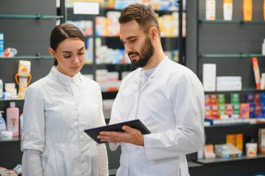 Eczanede iş birliği yapan eczacılar, çeşitli tıbbi ürünlerin rafları arasında bulunan dijital tablet üzerindeki envanter ve hasta bilgilerini kontrol ediyorlar.