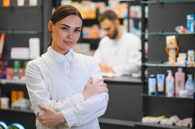 Gülümseyen kadın eczacı, kollarını kavuşturmuş, modern bir eczanenin arka planında çalışan bulanık bir erkek meslektaşla.
