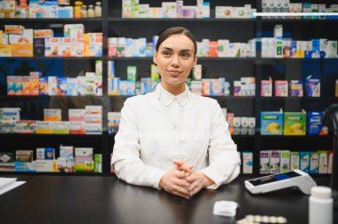 Dost canlısı kadın eczacı tezgahın arkasında duruyor, modern bir eczanede ilaç raflarıyla kameraya gülümsüyor.