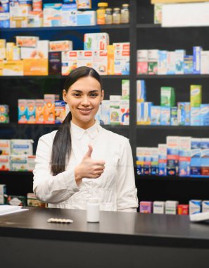 Mutlu genç bayan eczacı gülümsüyor, baş parmak hareketi yapıyor, eczane tezgahında ilaçlarla duruyor.