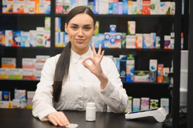 Eczanede duran genç bir kadın eczacı, elinde sağlık ve sağlık için ilaç tutarak iyi bir jest yapıyor.
