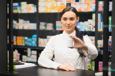 Genç bir kadın eczacı ya da doktor modern bir eczanenin tezgahında duran beyaz bir ilaç şişesini sunuyor.