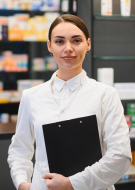 Beyaz önlüklü genç bir kadın elinde bir pano, arka planda ilaç raflarıyla modern bir eczanede duruyor.
