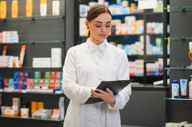 Üniformalı profesyonel kadın eczacı panoya yazı yazıyor, ilaç yönetiyor, modern bir eczanede sağlık ürünleri envanterini çıkarıyor.