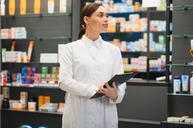 Modern eczane koridorunda duran eczacı, envanterleri kontrol ediyor ve sağlık ve ilaç ihtiyacı olan müşterilere yardım ediyor.