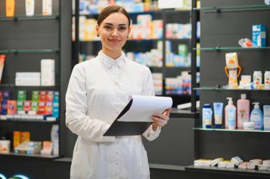 Elinde bir pano olan eczacı kadın, raflarında ilaç ve sağlık ürünleri olan modern bir eczanede duruyor. Profesyonel bakım sağlanıyor.