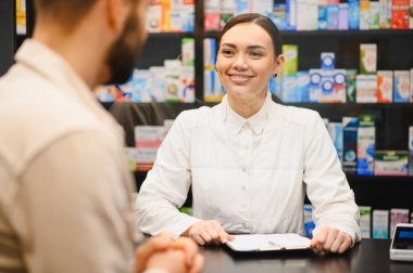 Dost canlısı bir kadın eczacı, gülümsüyor, kasada arka planda ilaç raflarıyla erkek bir müşteriyle konuşuyor.
