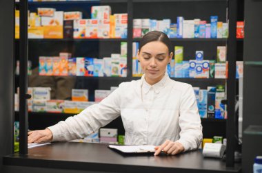 Eczane tezgahının arkasında duran kadın eczacı, belgeleri gözden geçiriyor ve hasta siparişlerini hazırlıyor.