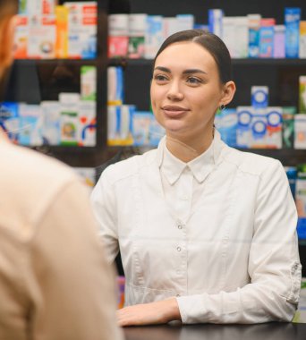 Beyaz önlüklü kadın eczacı müşteriyle konuşuyor, modern bir eczanede tıbbi danışmanlık ve hizmet veriyor.