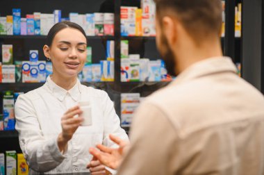 Eczacı elinde ilaç şişesiyle eczane raflarının önünde bir müşteriyle konuşuyor ve sağlık hizmeti sunuyor.