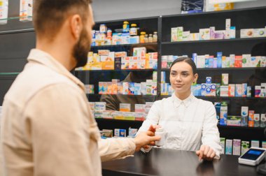 Tezgahın arkasında duran eczacı, modern bir eczane içinde erkek bir müşteriye bir şişe hap veriyor.