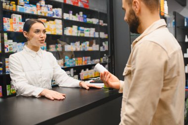 Tezgahta duran eczacı, erkek müşteriye alışverişinde yardım ediyor, profesyonel sağlık hizmeti sunuyor.
