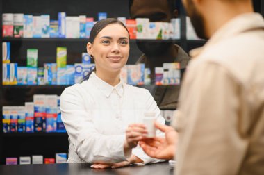 Gülümseyen eczacı tezgahta bir erkeğe ilaç veriyor, modern bir eczanede profesyonel sağlık hizmeti sunuyor.