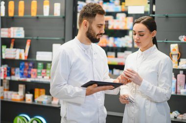 İki eczacı sağlık sorunlarını tartışıyor, modern bir eczanede uyuşturucu bilgileri ve su toplayan paketleri kontrol ediyorlar.