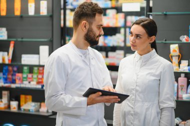 İki beyaz önlüklü eczacı modern bir eczanede dijital tablette envanter ya da reçete detaylarını tartışıyor.
