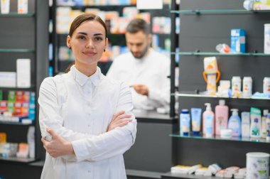 Gülümseyen kadın eczacı arka planda bulanık bir erkek arkadaşla modern bir eczanede kollarını kavuşturarak duruyor.