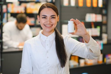 Beyaz önlüklü gülümseyen kadın eczacı boş bir ilaç şişesi gösteriyor, modern bir eczanede çalışıyor.