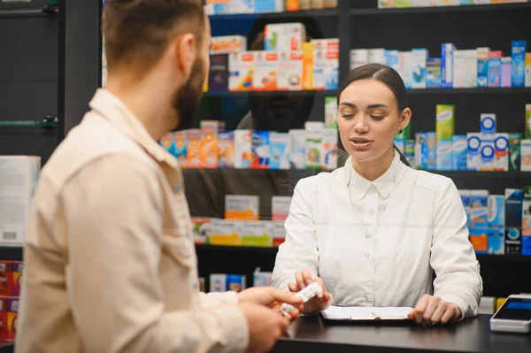 Eczanenin tezgahında duran eczacı elinde haplar olan bir erkek müşteriyle ilaçları tartışıyor.
