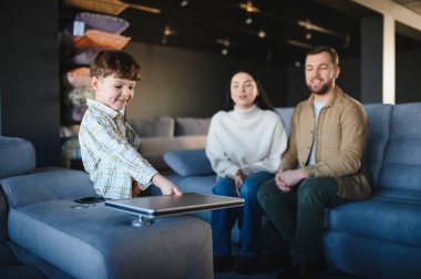 Genç bir aile, modern bir sergi salonunda yeni bir kanepeyi test ediyor. Oğulları rahat mobilya seçeneklerini incelerken ebeveynler gülümsüyor.
