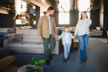 Ebeveynler ve oğulları el ele tutuşuyor, modern bir mobilya galerisinde yürüyor, yeni ev dekorasyonu arıyor.